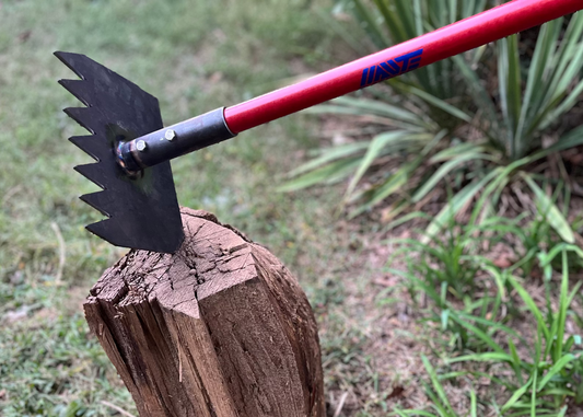 Trail tool with a red handle and black head on a wooden stump in a garden setting.